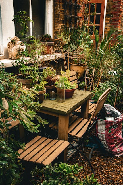 A small, cluttered garden patio situated next to a red-brick house with a wooden window frame and a glass-paneled door. The space features a weathered wooden outdoor table surrounded by matching woode